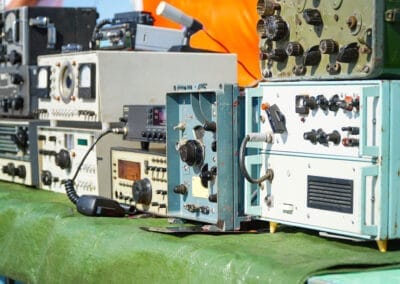 A collection of vintage radio and communication equipment is displayed, including radio transceivers, receivers, and amplifiers. The devices have various knobs, dials, and meters, and are arranged on a green tablecloth. The background is blurred.