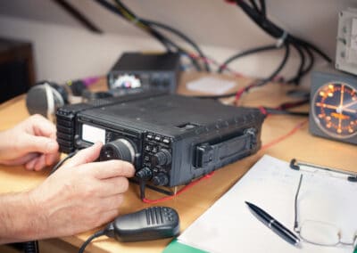 A person operates a ham radio transceiver on a cluttered desk. The transceiver is equipped with various knobs and buttons. Nearby, there is a microphone, pen, paper with notes, and an instrument displaying a radar-like screen with an orange circular display.