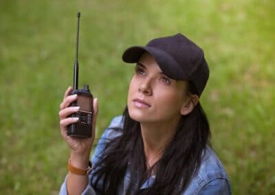 A woman wearing a black cap and denim shirt is outdoors holding a handheld radio, looking up with a focused expression. She is sitting on the grass, and the background is green and blurred.